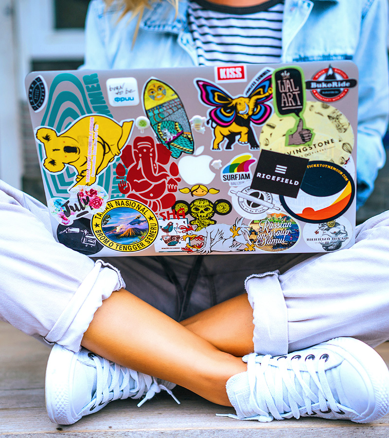 contact-laptop-stickers-img College student sitting cross-legged with a laptop covered in stickers on their lap